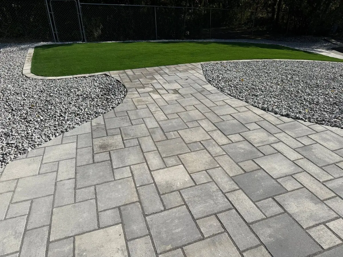 A wide grey paver patio laid in a herringbone pattern, curving between two crescent beds of grey landscape rock toward a bed of artificial turf held by a poured concrete curb, with a chain-link fence and trees behind
